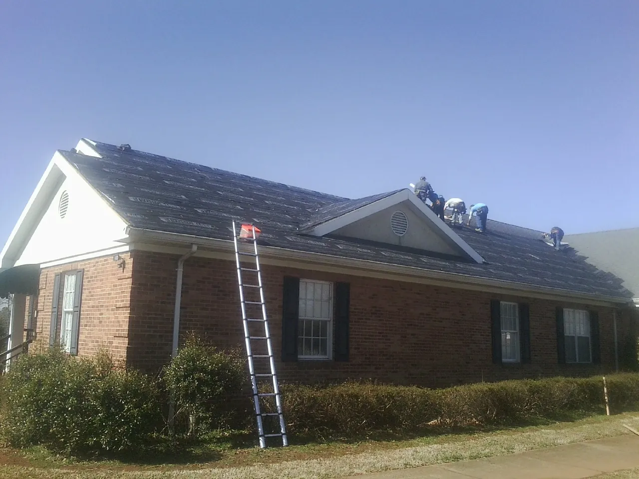 Crew tearing shingles off a brick ranch roof with a ladder set — Metal Roof Repair work by our crew in Leonardsville