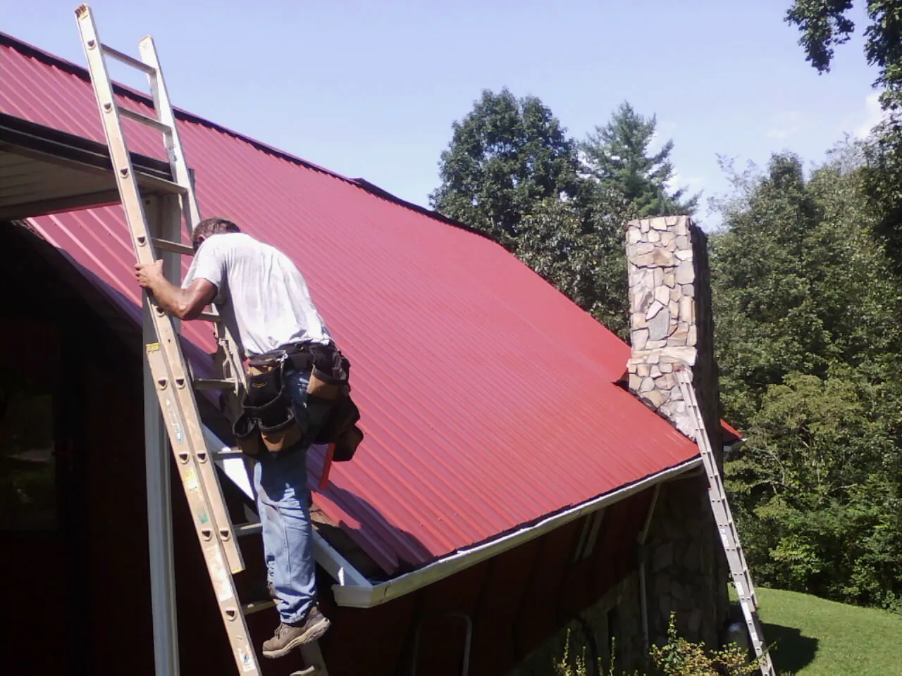 Roofer on a ladder beside a red metal roof and stone chimney — Soffit & Fascia Repair by Roofing Brothers in Leonardsville